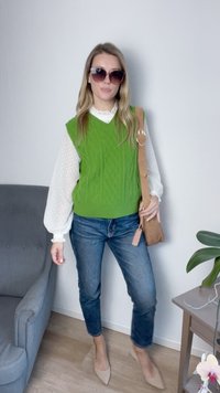 Colete de malha verde sobre uma blusa branca com mangas bufantes, combinado com jeans azuis e sapatos de bico fino bege. Bolsa de ombro castanha e óculos de sol.