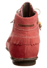 Kotníková bota z červeného semiše s ozdobnými třásněmi, koženými prvky a proužkovaným tkaným vnitřkem. Má zaoblenou špičku a plochou podrážku.
