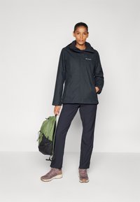 Black waterproof jacket with a hood, paired with dark pants and light brown hiking shoes; holding a green backpack with black mesh accents.