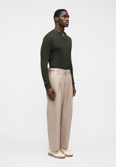 Άνδρας που στέκεται γυρισμένος προς τα δεξιά, φοράει σκούρο πράσινο ριπ πουλόβερ, ανοιχτό μπεζ πλισέ παντελόνι φαρδυάς γραμμής και λευκά αθλητικά παπούτσια με καφέ σόλες.