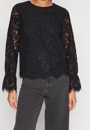 Musta pitsinen pitkähihainen pusero, jossa on kukkakuvio ja aaltoileva helma, yhdistetty tummanharmaisiin korkeavyötäröisiin farkkuihin.