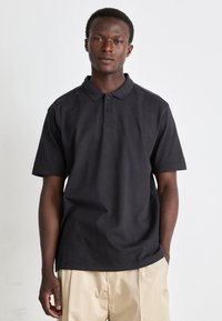 Polo negru din bumbac, cu guler, trei nasturi și un logo mic. Mâneci scurte și o croială lejeră, combinat cu pantaloni bej.