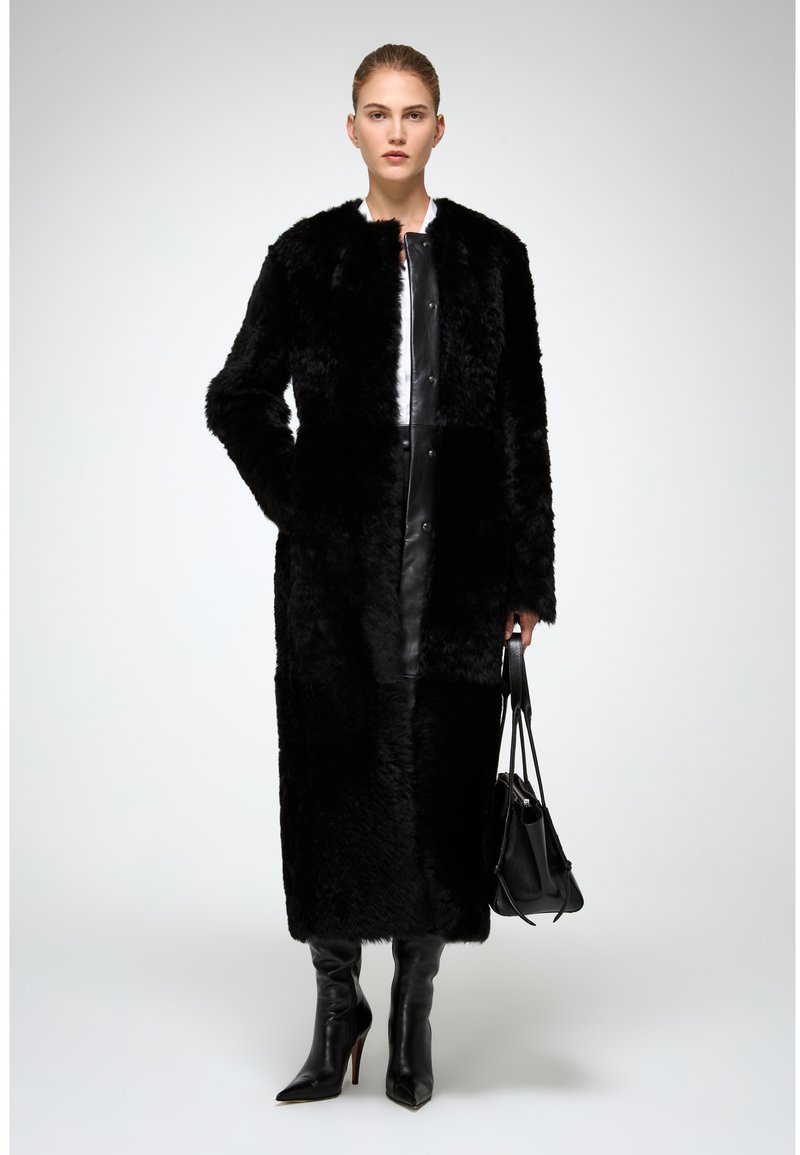 Cappotto in pelliccia sintetica nera con design senza colletto, maniche lunghe e bottoni frontali. Indossato sopra un vestito in pelle nera e abbinato a stivali con tacco a spillo neri e una borsa nera.