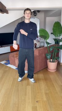 Črne jeans hlače in temno modra majica z dolgimi rokavi. Oseba stoji na lesenem tlaku ob kavču in velikih zelenih rastlinah v loncih.