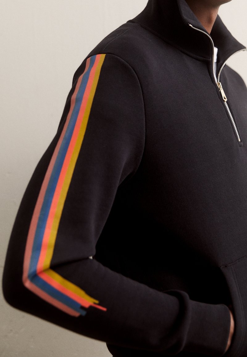 Uomo che indossa un felpa nera con zip e strisce multicolori lungo le maniche, con le mani in tasca, su uno sfondo chiaro e semplice.