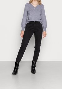 Grauer Strickpullover mit V-Ausschnitt, schwarze Skinny Jeans und schwarze Knöchelstiefel mit dicker Absätze, vor einem schlichten Hintergrund.