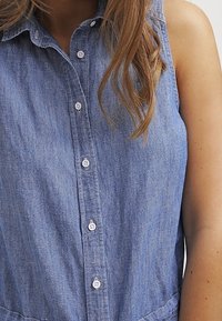 Ljusblå chambray ärmlös klänning med krage, knäppning fram och textilstruktur. Har en figurnära midja och utsvängd nederdel.