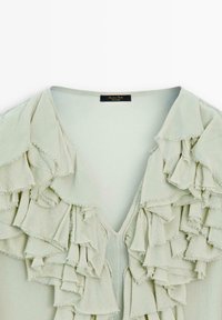 Jasnozielona bluzka z dekoltem w kształcie litery V i wieloma falującymi warstwami z przodu, uszyta z miękkiego, teksturowanego materiału. Metka widoczna w środku.