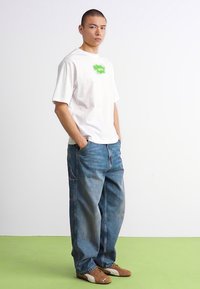Mann står med hendene i lommene, iført hvit grafisk T-skjorte, løse blå jeans og brune joggesko på grønn gulv.