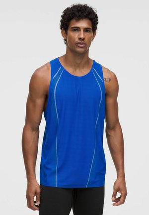 Mann mit lockigem Haar, der ein blaues ärmelloses Sporttop und schwarze Shorts trägt, vor einem schlichten hellen Hintergrund steht.
