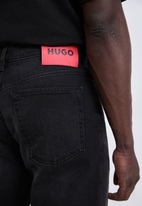 Jean en denim noir avec un label rouge HUGO bien visible à l'arrière, design à cinq poches standard et texture lisse.