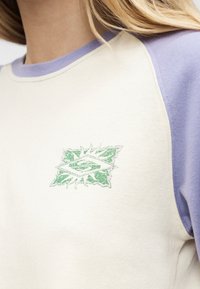 Κοντινό πλάνο ατόμου που φοράει μπλούζα ράγκλαν σε κρεμ και λιλά, με ένα μικρό πράσινο αφηρημένο έμβλημα στο στήθος.