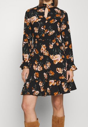 Femme portant une robe noire à fleurs avec manches longues, ourlet volanté, encolure fendue, et des bottes marron montant jusqu'aux genoux.