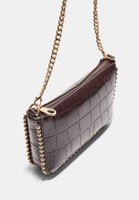 Torba iz krokodilje kože v globoki bordo rdeči barvi z zlatim verižnim trakom, zadrgo in dekorativnimi zlatimi zakovicami ob straneh.