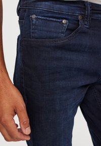 Tamni traperice s glatkom teksturom, s dva metalna gumba na prednjoj džepu i minimalnim detaljima šivanja.