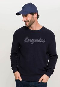 Tums zila adīta džemperis ar gaiši pelēku izšuvumu "bugatti". Saskaņots ar atbilstošu tums zilu cepuri ar logotipu.