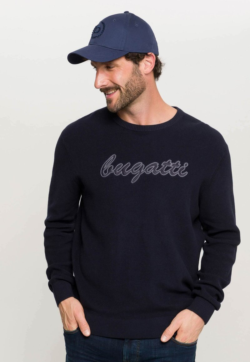 Maglione lavorato a maglia blu navy con "bugatti" ricamato in grigio chiaro. Abbinato a un cappello blu navy coordinato con logo.
