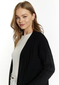 Fekete kötött kardigán, kábelmintával, világosszürke felsőre húzva. Hosszú haj, laza hullámokkal, színes, lógó fülbevalókkal.