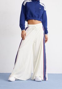 Felpa blu scuro con strisce bianche, abbinata a pantaloni bianchi a gamba larga con texture a coste e strisce laterali rosse e blu. Sneakers bianche.