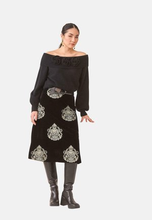 Mujer con un suéter negro hombros caídos con detalles florales, una falda negra con patrones beige intrincados, un cinturón y botas negras hasta la rodilla.