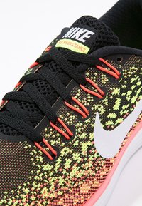 Černé tenisky s texturovaným svrškem, který má neonový vzor. Bílé logo Nike ve tvaru swoosh, růžová podrážka a černé tkaničky s oranžovými akcenty.