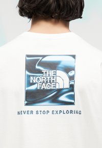 Άτομο που φοράει λευκό πουκάμισο με μπλε μεταλλικό λογότυπο The North Face και το κείμενο «Ποτέ μην σταματάς να εξερευνάς» στην πλάτη.