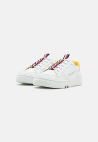 Sneaker bianche con dettagli gialli, tomaia in pelle con texture, lacci a righe rossi, bianchi e blu, e una spessa suola in gomma bianca.