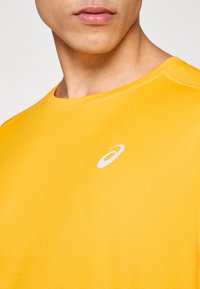 Κίτρινο αθλητικό πουκάμισο με στρογγυλή λαιμόκοψη, κατασκευασμένο από υφή ύφασμα. Διαθέτει μικρό λευκό λογότυπο στην αριστερή πλευρ ά του στήθους.