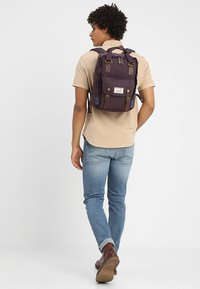 Dark purple backpack with brown leather accents and buckle straps, worn on the back of a person in a beige shirt and blue jeans.