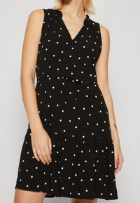 Robe noire sans manches à pois blancs, encolure en V, épaules froncées, taille élastique et ourlet à volants. Texture de tissu lisse.