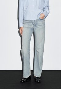 Jeans in denim blu chiaro con taglio dritto, caratterizzati da una leggera scoloritura e design classico a cinque tasche, abbinati a una camicia a righe blu chiaro e scarpe nere.