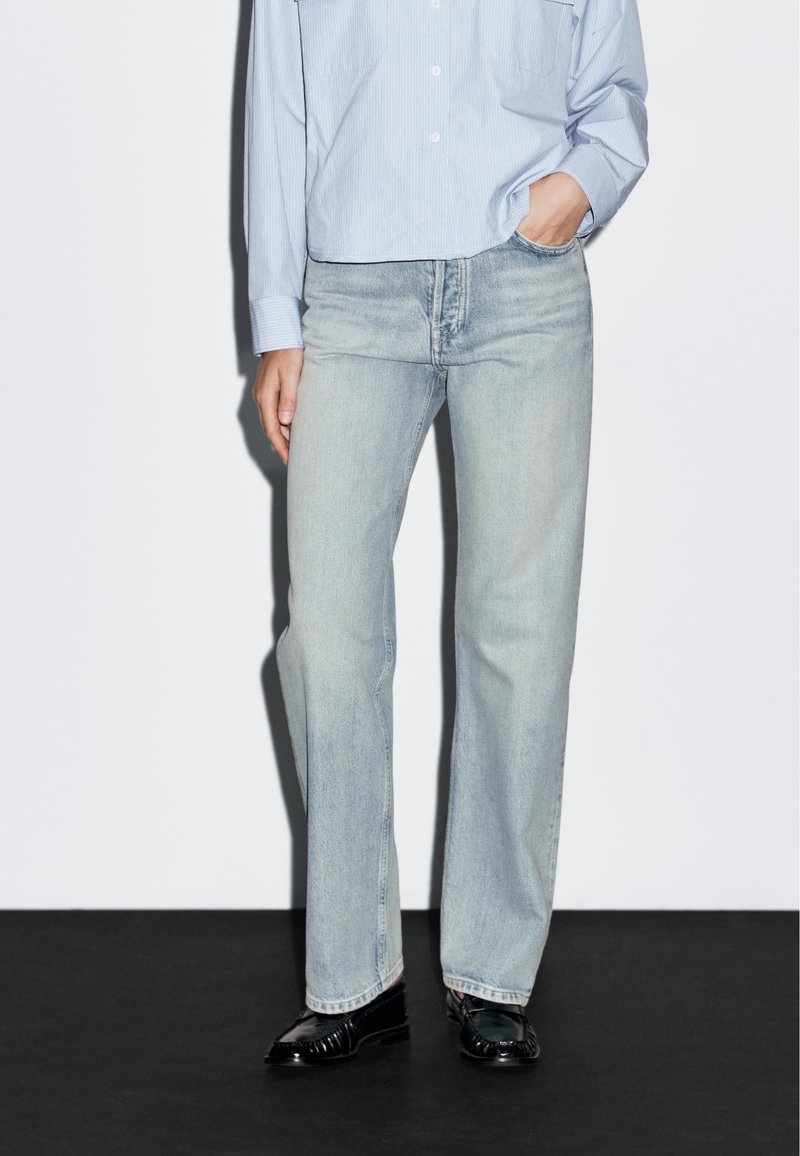 Jeans in denim blu chiaro con taglio dritto, caratterizzati da una leggera scoloritura e design classico a cinque tasche, abbinati a una camicia a righe blu chiaro e scarpe nere.