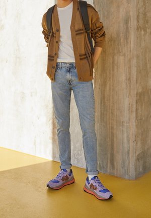 Person trägt hellblaue Jeans, beige-braune Strickjacke, weißes Hemd, lila-orange Sneaker und schwarzen Rucksack und steht vor einer strukturierten Wand.