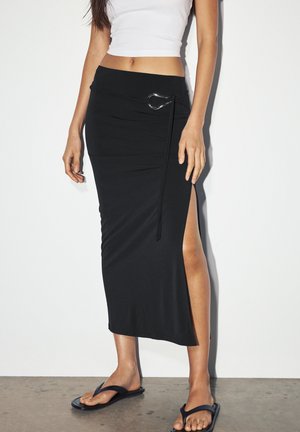 Zwarte, aansluitende midi-rok met een zij-split, geaccentueerd door een zilveren D-ring detail en een zachte, rekbare stof. Gecombineerd met zwarte slippers.