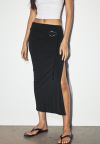 Black, form-fitting midi skirt with a side slit, accentuated by a silver D-ring detail and smooth, stretchy fabric. Paired with black flip-flops.