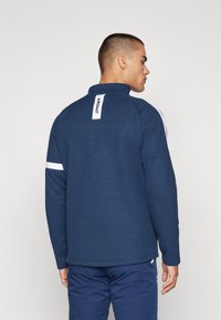 Pull-over bleu marine avec un col montant, en tissu texturé, et des bandes blanches sur les manches. Dispose d'un onglet logo au dos du col.