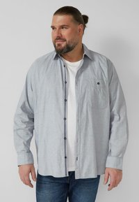 Lichtgrijs overhemd met knopen en een subtiel ruitpatroon, zwarte knopen en een borstzak, gedragen over een wit T-shirt.