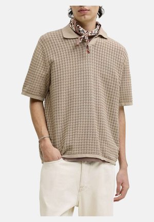 Bărbat purtând un polo tricotat bej cu mâneci scurte, pantaloni albi cu o mână în buzunar și un fular cu model legat în jurul gâtului.