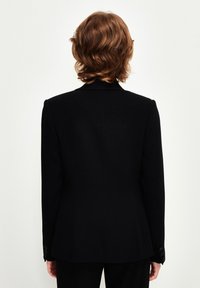 Blazer nero con spalle strutturate, texture del tessuto, spacco posteriore singolo e polsini con bottoni, visto da dietro.