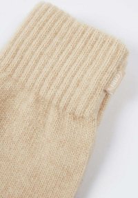 Chaussette beige en laine tricotée avec bord-côte à la cheville et petite étiquette en tissu sur le côté, posée à plat sur une surface blanche.