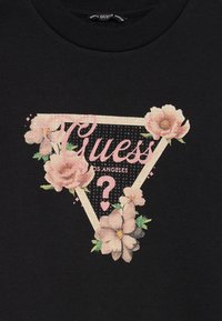 Črn material s rožnatim cvetličnim trikotnikom, z notacijo "Guess" in "Los Angeles" znotraj trikotnika.