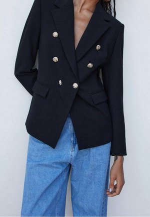 Personne portant un blazer noir croisé à six boutons dorés et un jean bleu taille haute, debout devant un fond uni clair.
