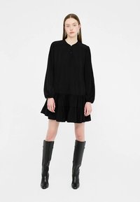 Sort langærmet kjole med plisseret tekstur, knaplukning ved halsen og lagdelt flæsekant; stylet med knæhøje sorte støvler.