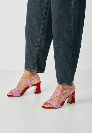 Lyserøde ruskinds sandaletter med brede stropper og åben tå, med en rød blokkadelhæl. Justerbar spænde ved anklen til lukning.