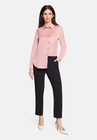 Jasnoróżowa koszula na guziki z długimi rękawami, spiczastym kołnierzem i zaokrąglonym, przedłużonym dołem, zestawiona z czarnymi dopasowanymi spodniami i białymi szpilkami.