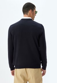 Granatowy sweter robiony na drutach z długimi rękawami i białym kołnierzykiem, z prążkowanym dołem i mankietami. Model nosi okulary przeciwsłoneczne i khaki spodnie.