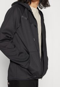 Sudadera con cremallera negra hecha de una tela suave, con capucha ajustable, dos bolsillos frontales y sutiles acentos a rayas en los hombros.