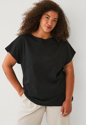 Krøllet kvinde iført en løs sort kortærmet skjorte og lyse beige bukser, stående med den ene hånd i lommen mod en ensfarvet baggrund.