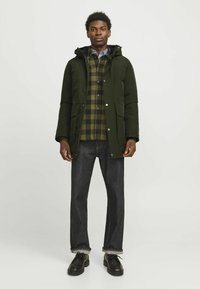 Roheline park kleit koos kapuutsiga, millel on kaks esist taskut, kantud ruudulise särgi peal, koos tumedate teksade ja mustade nööpidega kingadega.