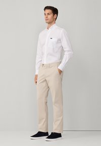 Camicia bianca con bottoni e tasca sul petto, pantaloni beige e sneakers nere. Tessuto morbido, vestibilità sartoriale e design semplice.